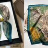 A teal and olive green floral print silk scarf folded in a square frame.