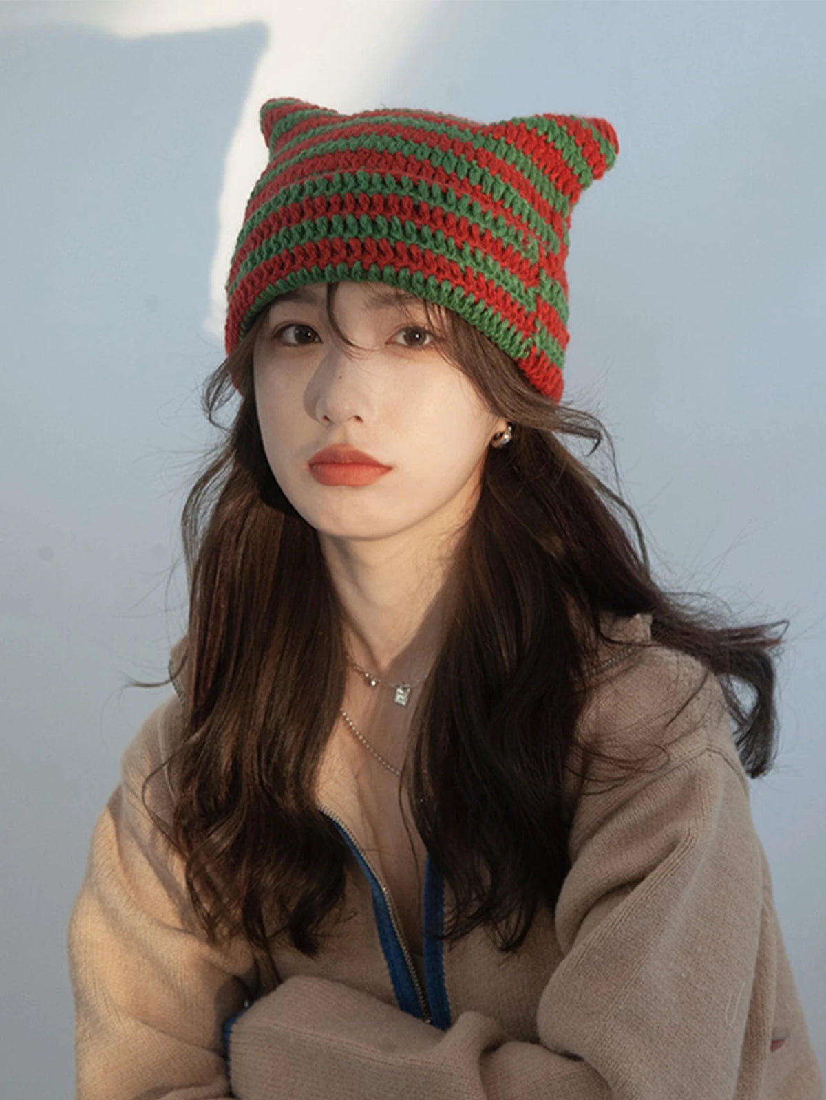 A woman wearing a green and red crocheted cat ears beanie hat and a beige sweater.