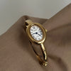 A gold-toned quartz analog watch with an oval face and Roman numerals is displayed on a beige fabric.