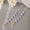 A delicate pearl beaded eyewear chain featuring lavender beads and gold accents displayed on a gray background.