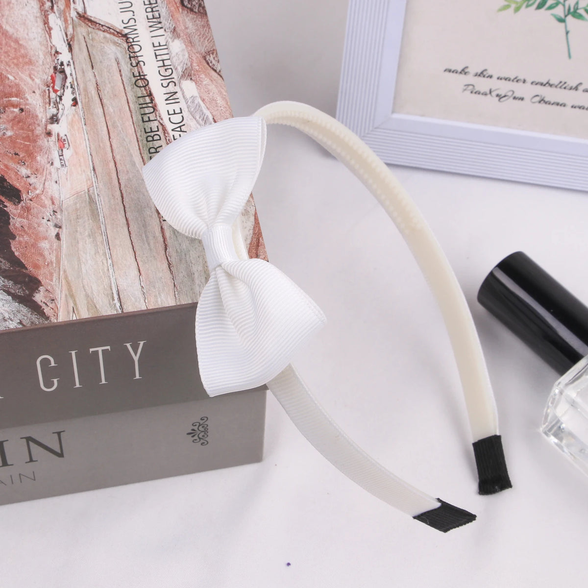 A white grosgrain ribbon hair bow is displayed next to a book and a black cosmetic product.