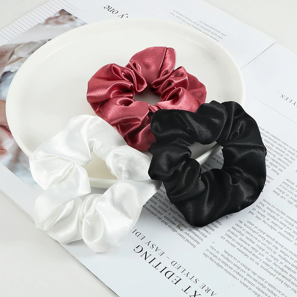 Three satin scrunchies in shades of pink, black, and white are displayed on a white plate.