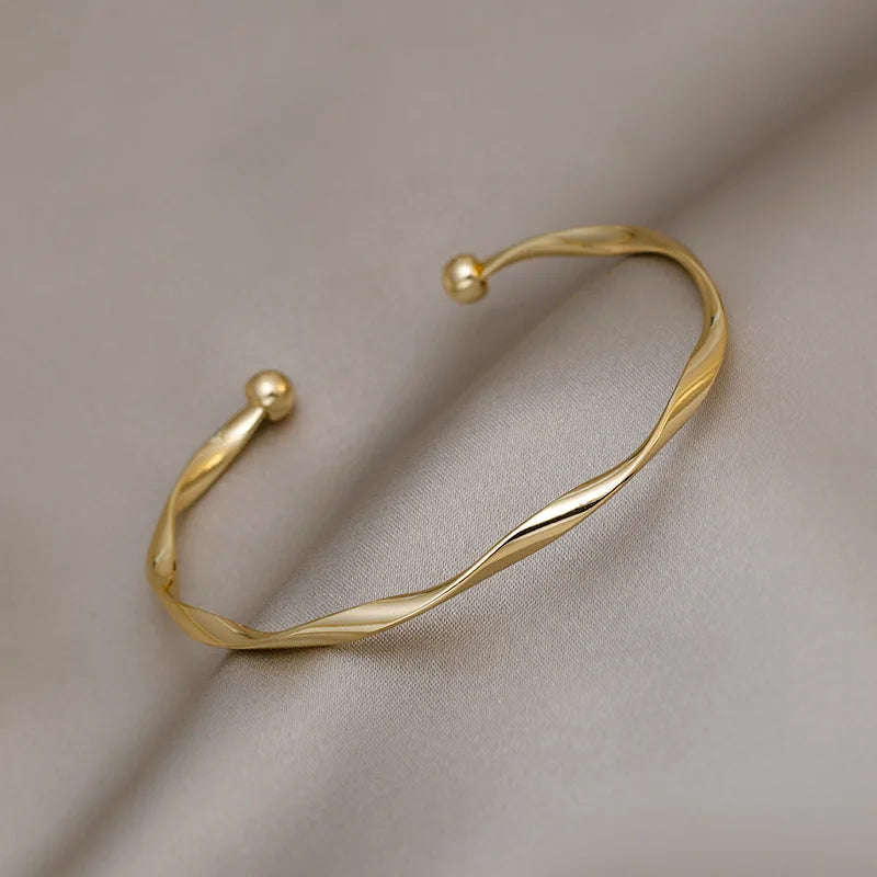 A minimalist gold-tone twisted cuff bangle resting on a neutral gray fabric.