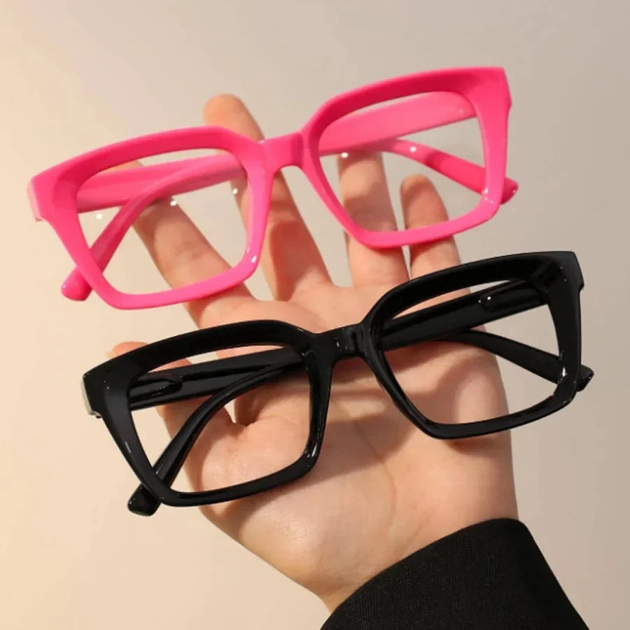 A hand holds a pair of pink and black square reading glasses against a light background.