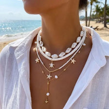 Gros plan d'une femme portant un collier superposé avec des charmés coquillages et étoiles de mer sur fond de plage.