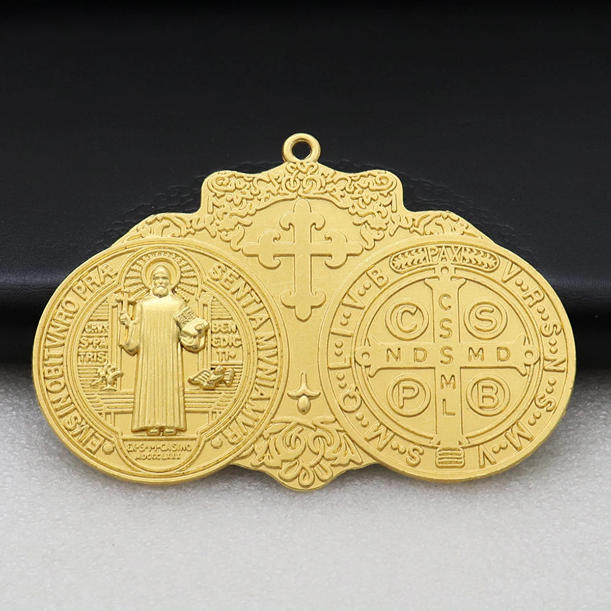 A gold-colored Saint Benedict protection medal with a cross and religious symbols, displayed on a dark background.