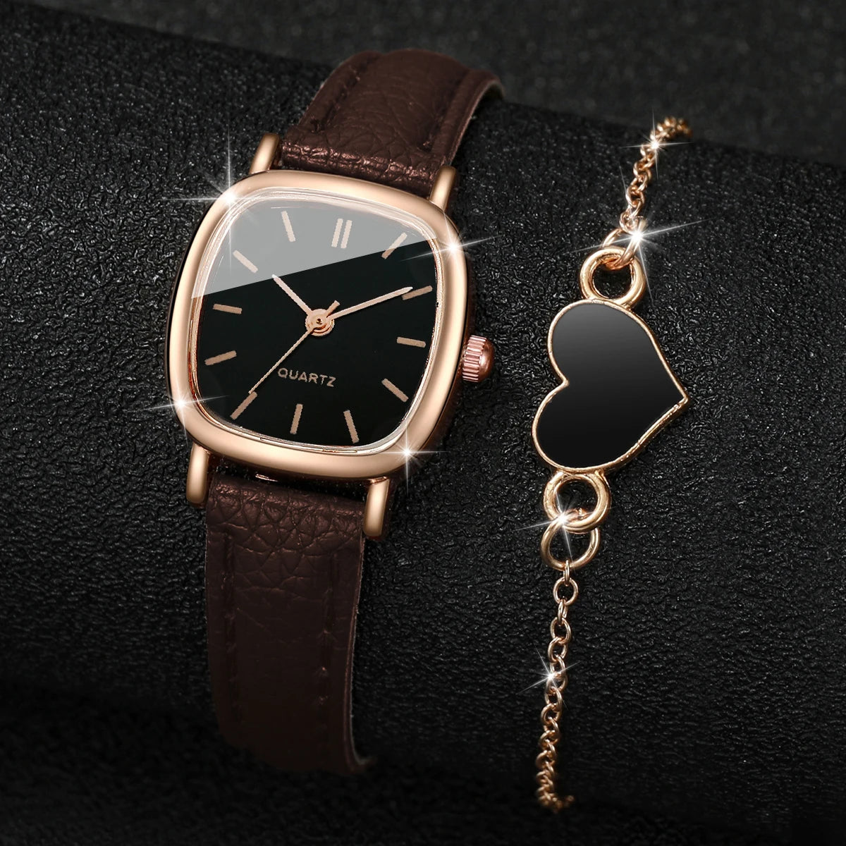 A square gold-toned watch with a dark face and brown leather strap next to a heart-shaped gold chain pendant.