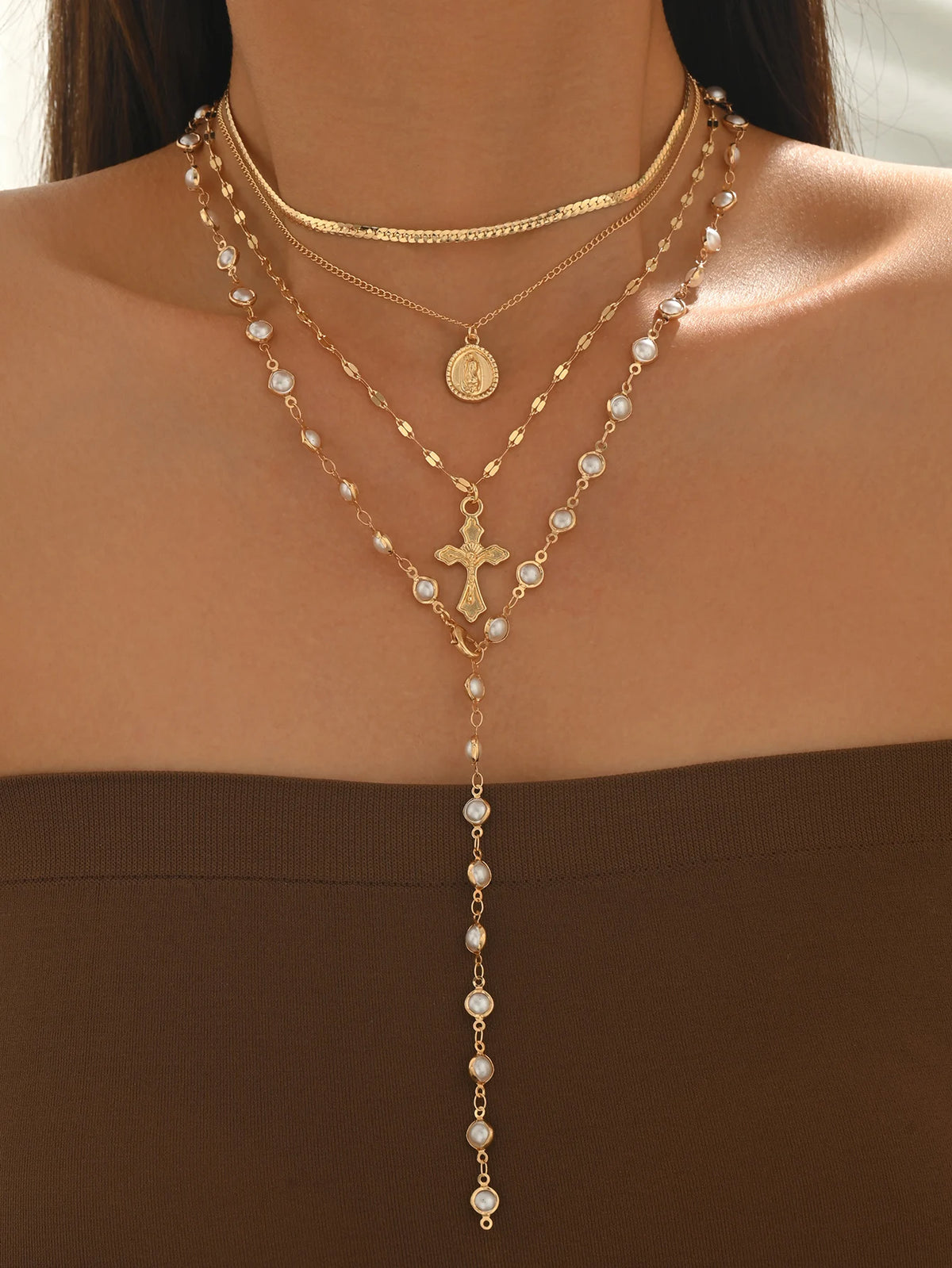 A close-up of a layered gold necklace featuring pearl beads and a cross pendant worn against a brown top.