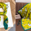 A bright yellow silk scarf with a floral print and green trim is displayed on a brown leather sofa.