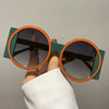 A hand holds a pair of round double color fashion sunglasses with green frames and orange accents.