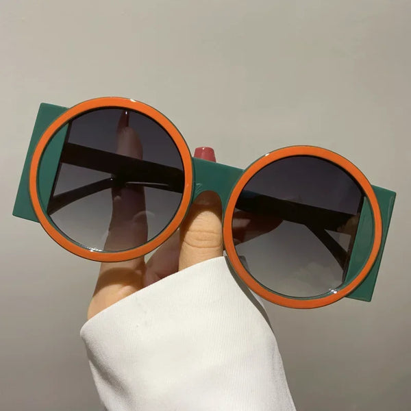 A hand holds a pair of round double color fashion sunglasses with green frames and orange accents.