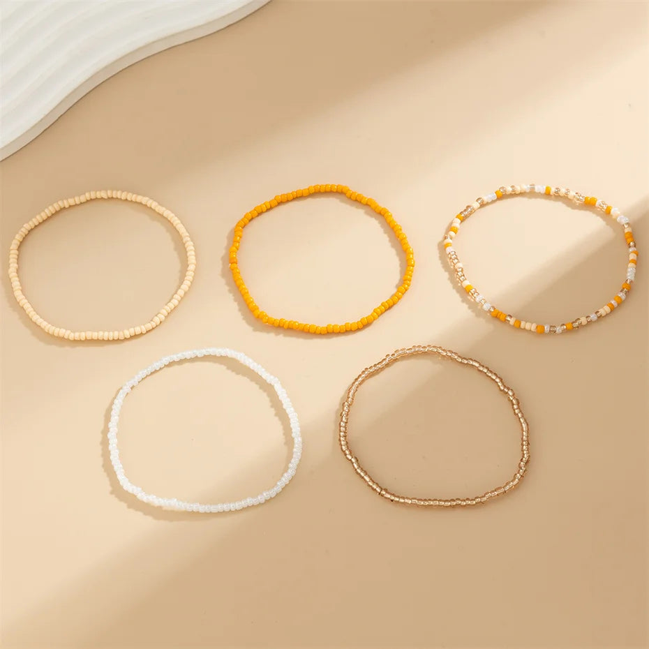 A collection of five beaded bracelets and anklets in various shades of yellow, white, and orange laid on a beige surface.