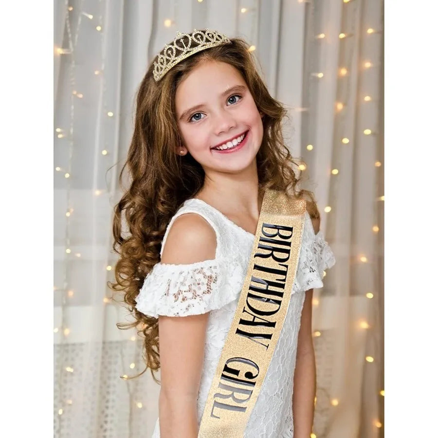 A smiling girl wearing a gold birthday tiara crown and sash against a soft, light background.