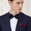A man wearing a navy suit jacket with a white shirt and black knit bow tie and a maroon pocket square.
