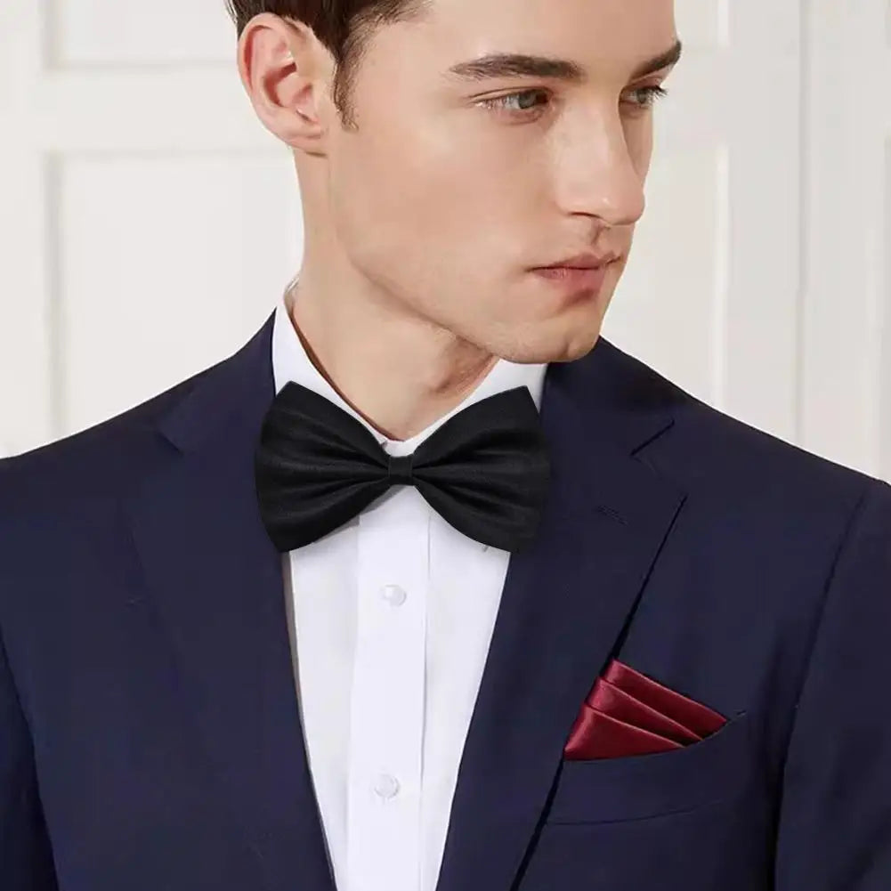 A man wearing a navy suit jacket with a white shirt and black knit bow tie and a maroon pocket square.