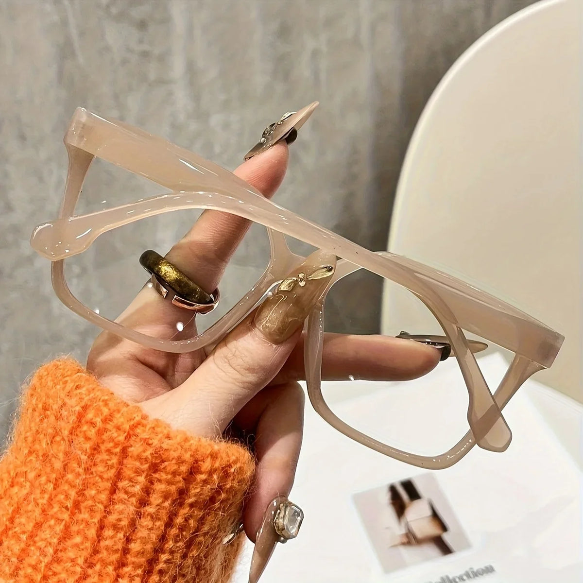 A hand holds a pair of elegant geometric reading glasses with transparent lenses against a light background.