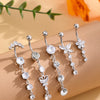 A set of five gold stainless steel dangled belly button rings with clear stones displayed on a beige surface.