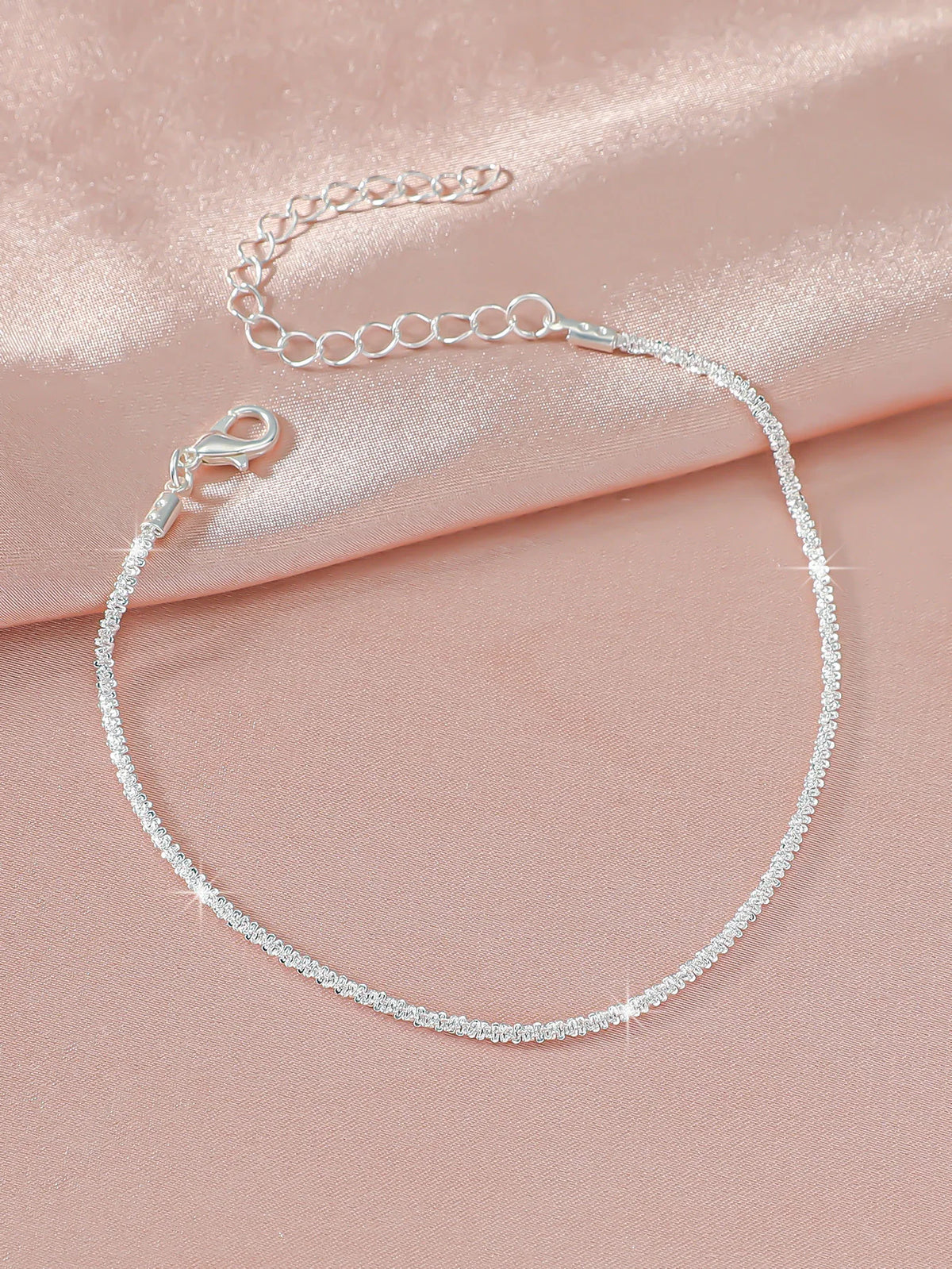 A delicate sparkling zircon heart cross anklet laid on a soft pink satin fabric.