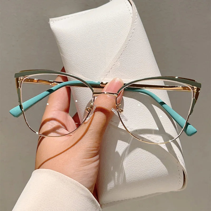 Una mano sostiene unas gafas de computadora con diseño de gato metálicas y marcos azul claro contra una libreta de cuero blan