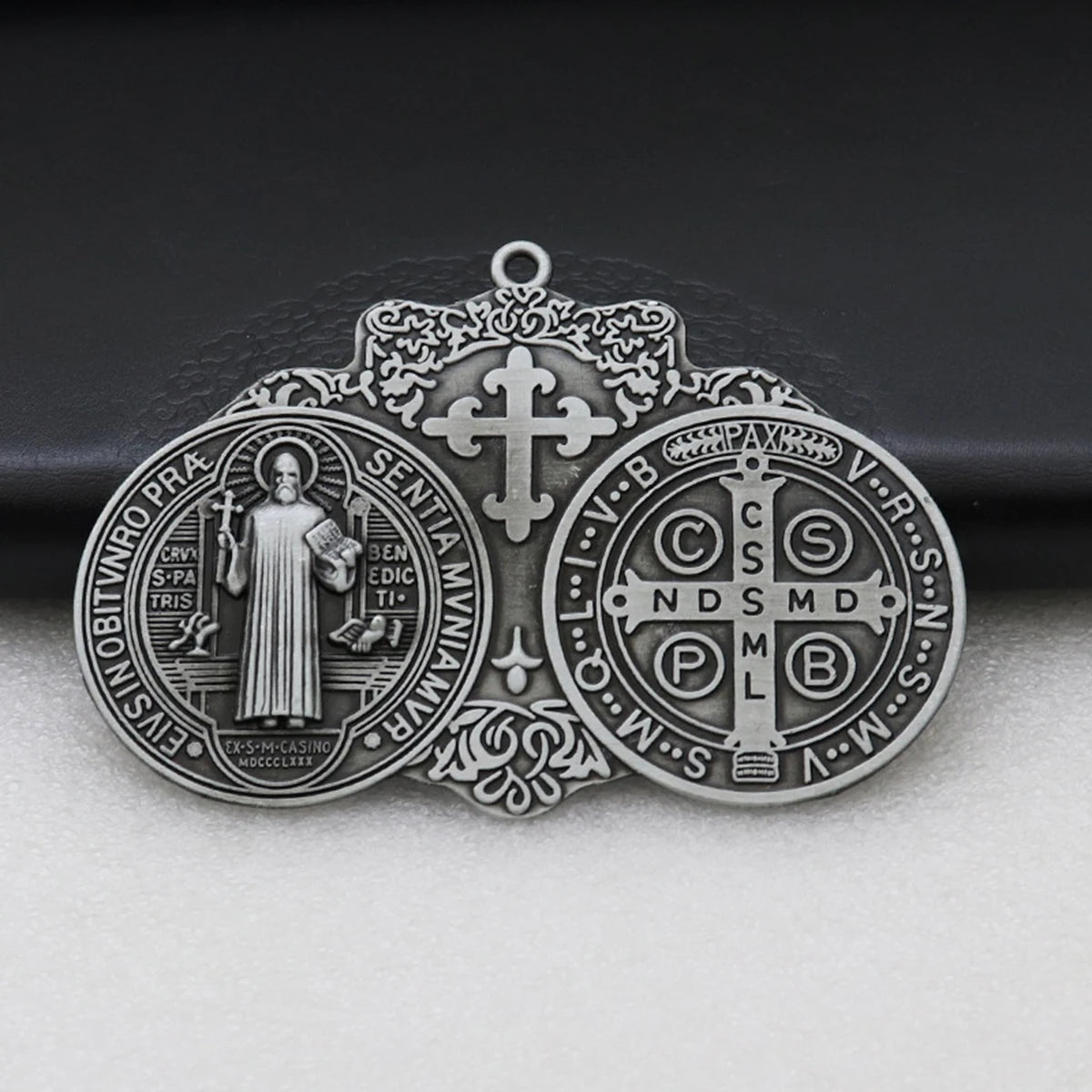 A silver Saint Benedict protection medal featuring a cross and various religious symbols, displayed on a dark surface.