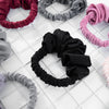 A collection of heatless hair curling scrunchies in various shades of gray, pink, and black arranged on a white grid.