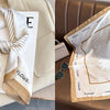A beige silk scarf with a floral print and gold trim is displayed on a light-colored surface.
