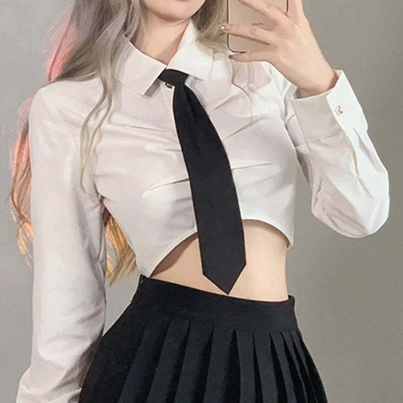 A woman wearing a white cropped shirt, black tie, and black pleated skirt.
