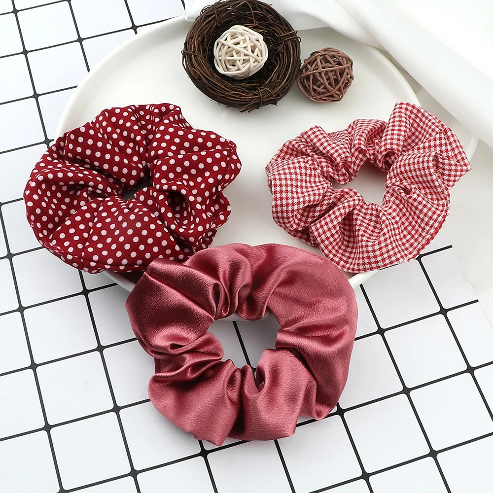 A display of three fabric scrunchies, including a red polka dot, a red and white checkered, and a satin pink one, alongside a