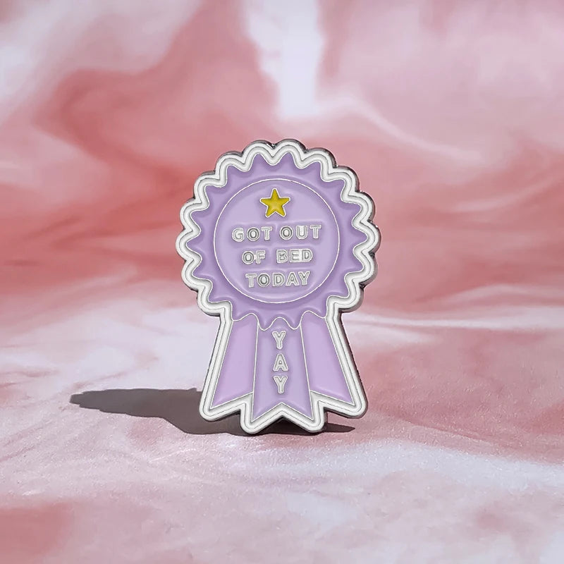 A purple enamel pin shaped like a ribbon award with the text Got Out Of Bed Today and a star.