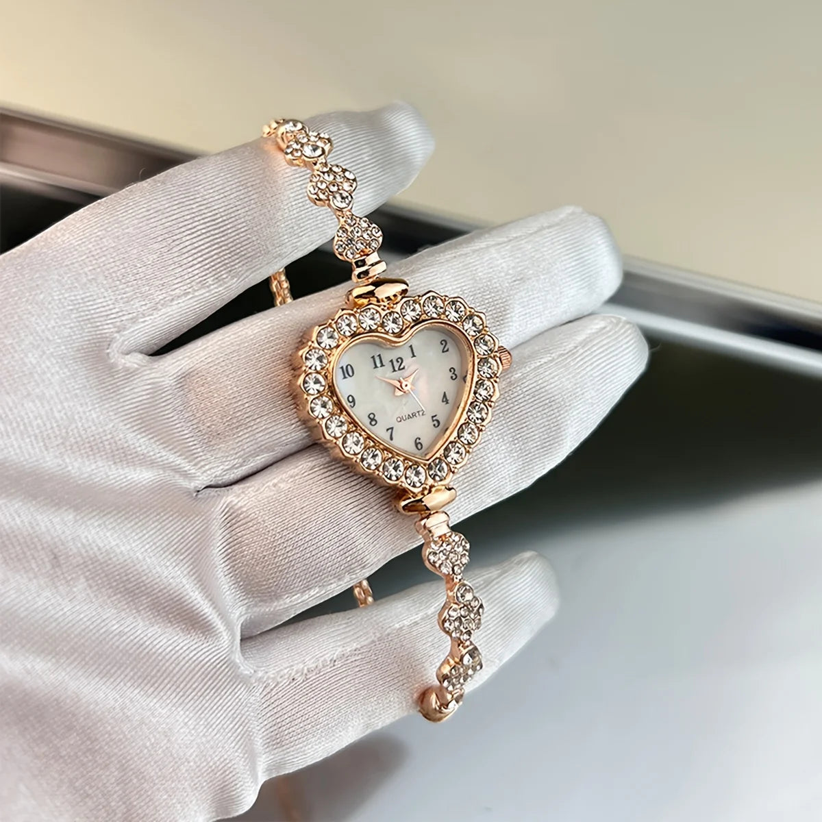 A hand wearing a light pink glove holds a sparkling heart rhinestone quartz watch.