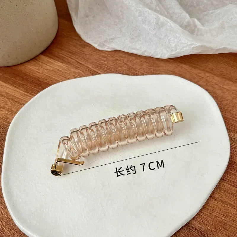 A clear rubber braided ponytail holder with a gold accent is displayed on a white ceramic plate.