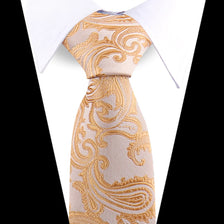 A close-up of an elegant men's floral paisley silk necktie in beige with gold embroidery, shown against a white shirt.