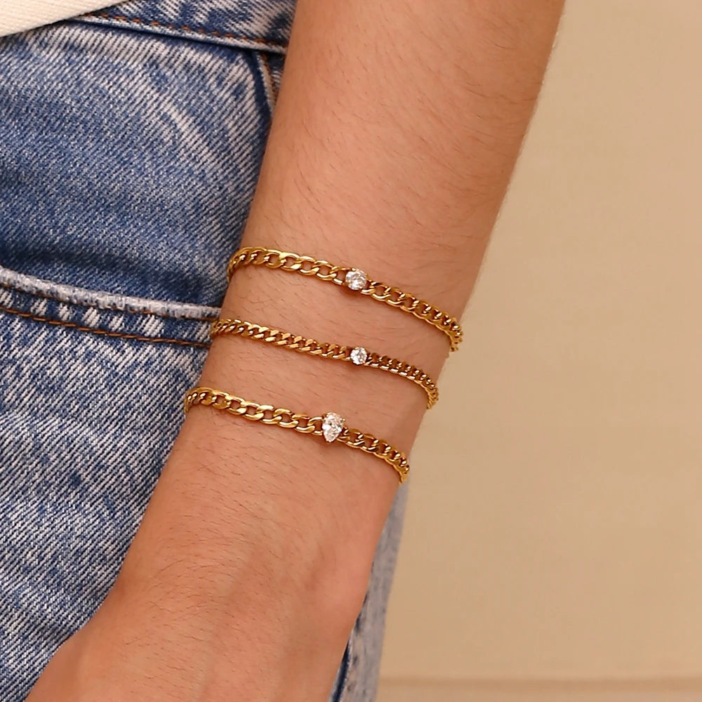 A close-up of a hand wearing three gold-toned charm bracelets with round water drop zircon stones on a denim background.