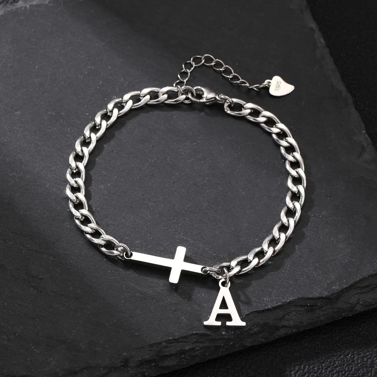 A silver stainless steel Cuban chain bracelet with a cross and the letter A charm resting on a black surface.