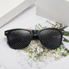 A pair of black vision correction pinhole glasses resting on a white surface with greenery.