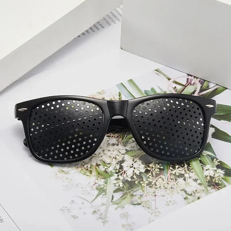 A pair of black vision correction pinhole glasses resting on a white surface with greenery.