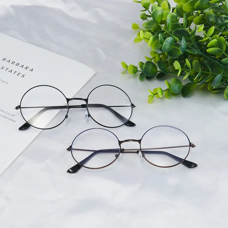 Un par de gafas redondas de metal anti-luz azul sobre una superficie blanca junto a follaje verde.