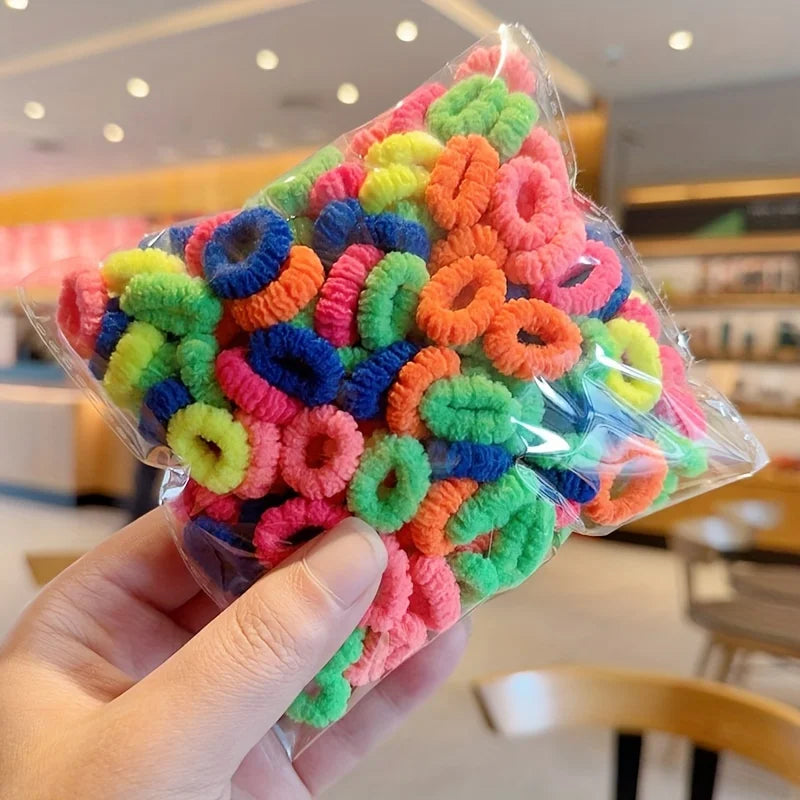 A hand holds a clear plastic bag filled with colorful, fuzzy elastic hair bands in various shades of pink, orange, yellow, an