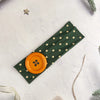 A dark olive green fabric headband with small gold dots and an orange circular accent on a white background.