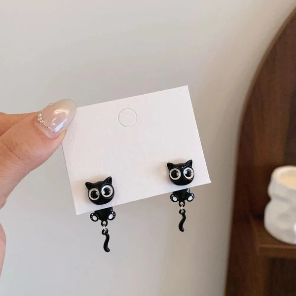 A pair of black cat stud earrings with dangling charms displayed on a white card against a light background.