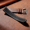 A black leather watch strap with a silver buckle resting on a brown textured surface.