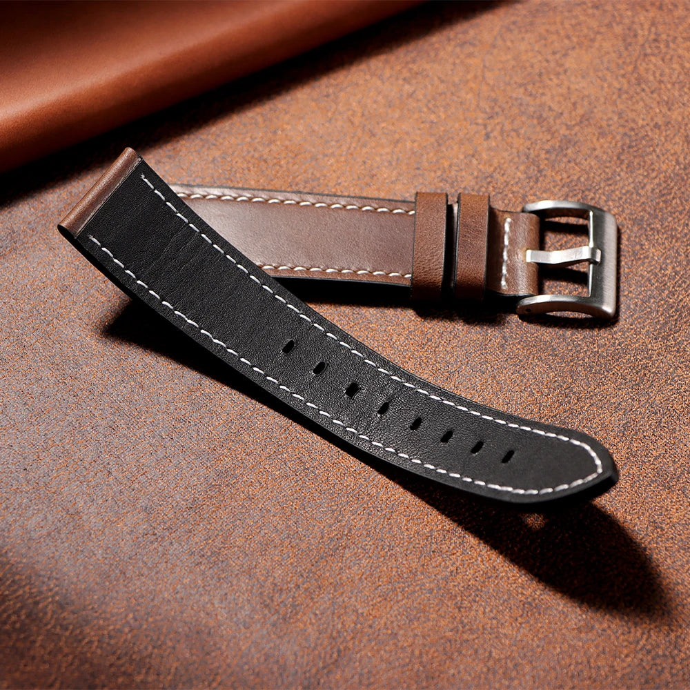 A black leather watch strap with a silver buckle resting on a brown textured surface.