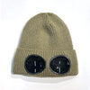 A close-up of a olive green knit windproof hat with round black goggles attached to the front.