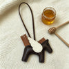 A dark brown horse pendant charm with white wings and a leather cord rests on a cream-colored fabric next to honey and a wood