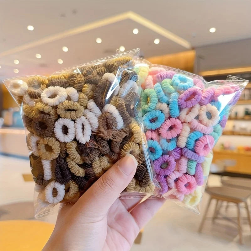 A hand holds a clear plastic package filled with colorful elastic hair bands for girls.