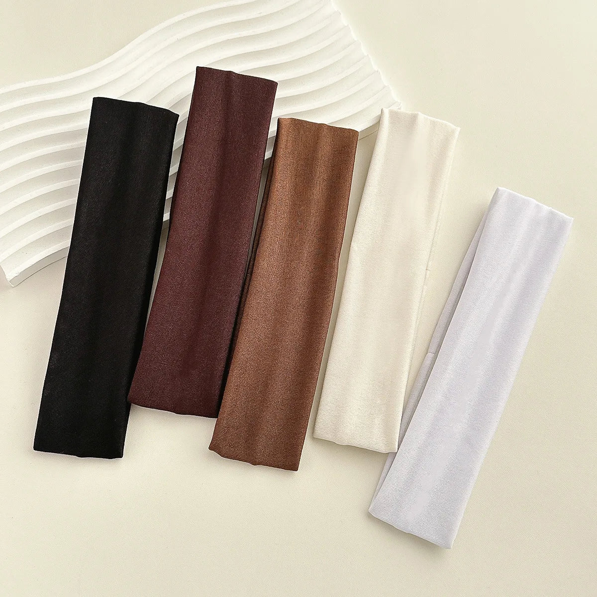 A collection of five solid fabric headbands in shades of black, brown, beige, and light gray are displayed on a light backgro