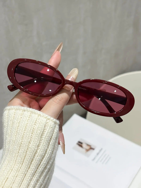 A hand holds a pair of maroon cat eye sunglasses against a light background.