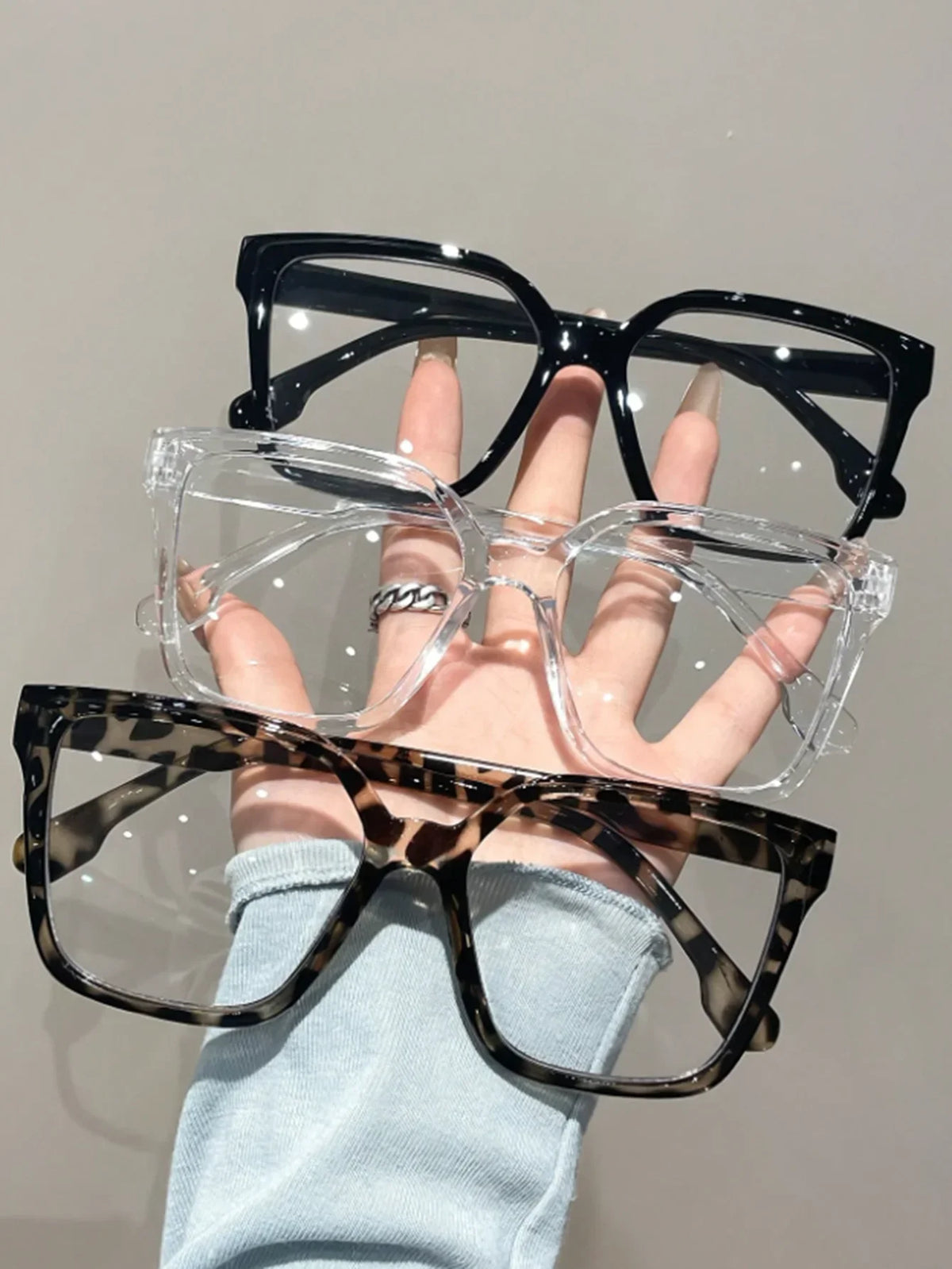 Una mano sostiene unas gafas cuadradas transparentes con montura negra y un par de gafas de estampado de leopardo.