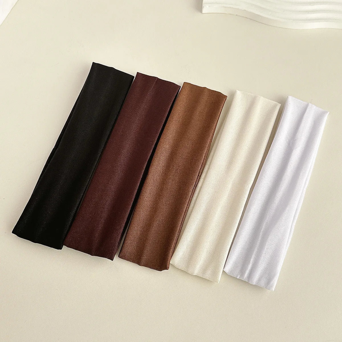 Five solid colored fabric headbands in black, brown, tan, cream, and white laid out on a light surface.