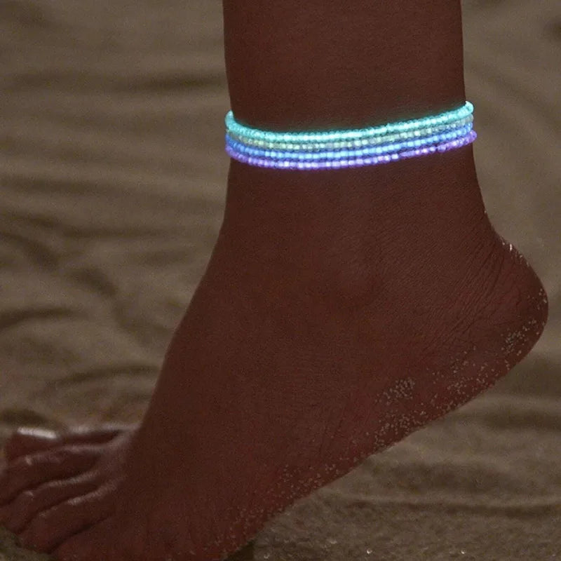 A close-up of a woman's ankle wearing luminous glow-in-the-dark beaded anklets that emit a blue and purple light.